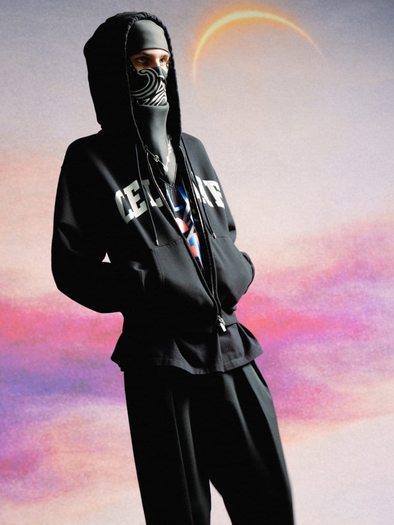 Model wearing a black hooded jacket and patterned face covering against a painted sunset sky background.