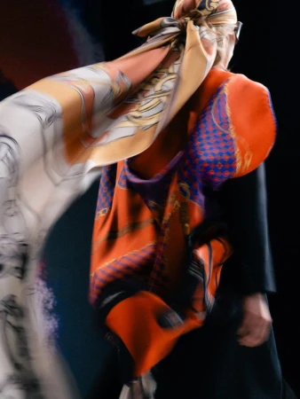 Rear view of a model walking in a flowing printed silk headscarf and orange patterned drape over a black tailored coat.