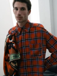 Man wearing an orange plaid shirt with a patterned scarf draped over his shoulder against a plain wall.