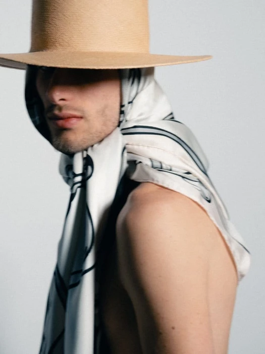 Man wearing a wide-brim tan hat and a draped printed silk scarf over bare shoulders against a white studio background.