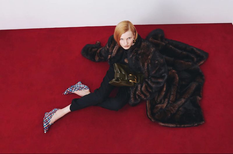Fashion editorial shot of a redhead woman lying on a red surface wearing a fur coat, black pants, and patterned high heels, holding a clutch.