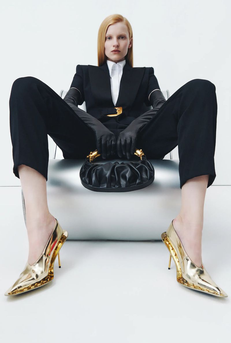 Fashion shoot featuring a woman in a black suit, gold high heels, holding a black purse, seated against a studio backdrop.