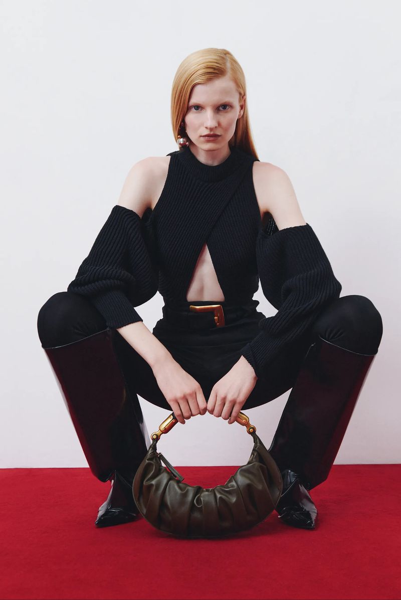 A slender, fair-skinned adult female model in a striking black cutout sweater, black pants, and black knee-high boots poses with a green purse.