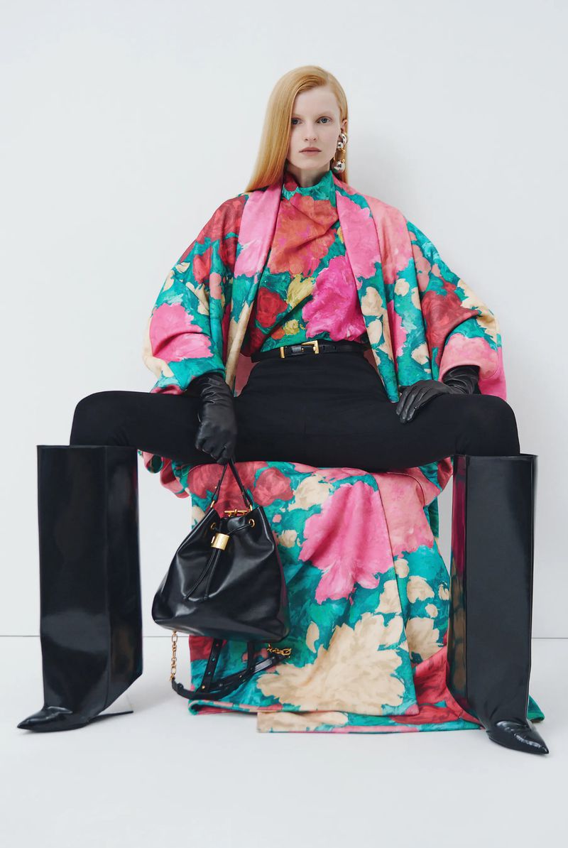 Fashion model in studio wears floral jacket, black pants, and patent leather boots, showcasing fashion-forward editorial style.