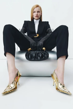 Model seated on a silver chair in a black tailored suit, black leather gloves, and metallic gold pointed pumps holding a ruched handbag.