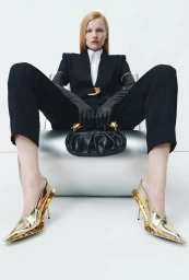 Model seated on a silver chair in a black tailored suit, black leather gloves, and metallic gold pointed pumps holding a ruched handbag.