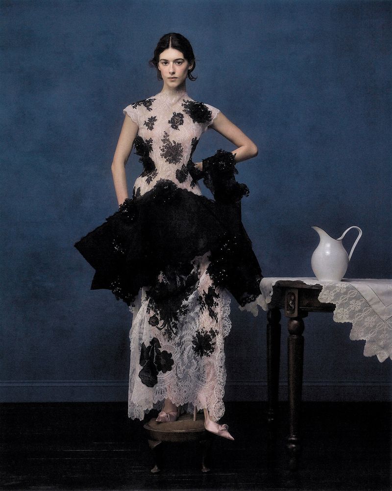 Daisy Edgar-Jones in an ornate black and pale lace gown standing beside a table with a white pitcher against a deep blue background.