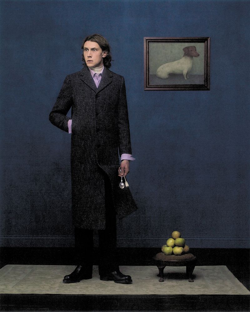 George MacKay in a long charcoal coat standing beside a blue wall with a framed dog painting and a stack of green apples.