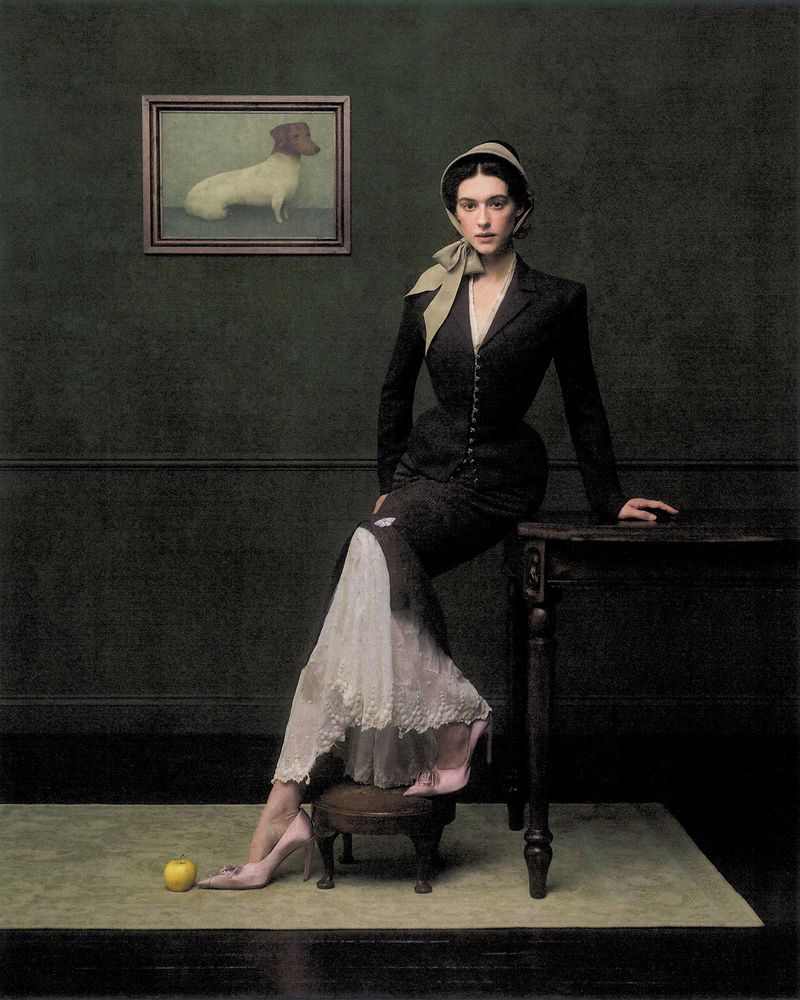 Woman in dark vintage-style jacket and bonnet seated beside a dark table against a deep green wall with a dog painting