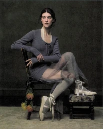 Daisy Edgar-Jones seated on a vintage chair wearing a grey knit top, sheer tulle skirt, and ribboned ballet shoes against a dark wall.