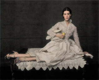 Daisy Edgar-Jones in an off-white lace dress reclining on a lace-draped bench against a dark textured backdrop holding a yellow fruit.