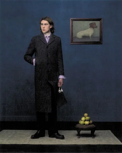George MacKay in a long charcoal coat standing beside a blue wall with a framed dog painting and a stack of green apples.
