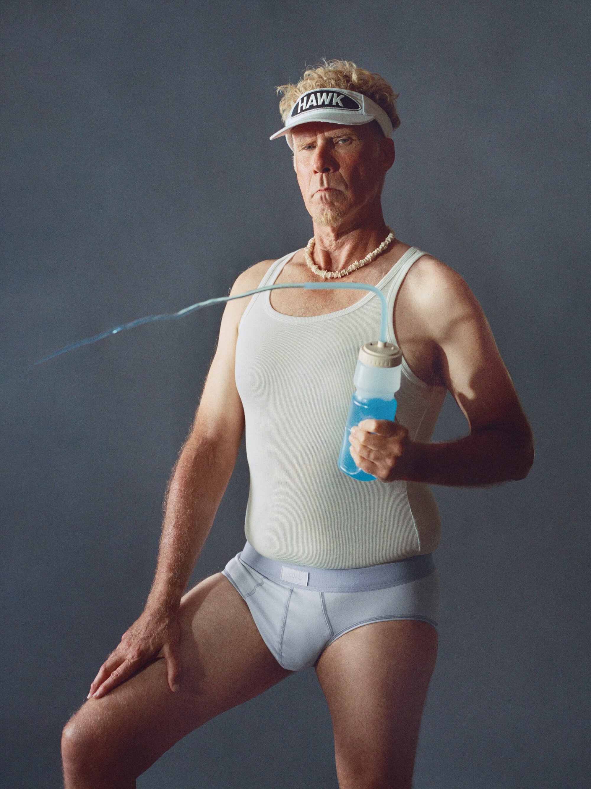Will Ferrell wearing a white tank top, grey underwear, visor, and shell necklace holding a blue water bottle against a grey backdrop.