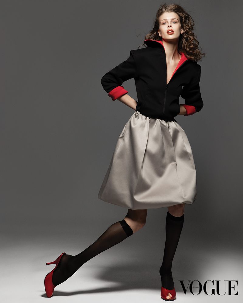 Model wearing a black and red jacket with a grey skirt and red heels against a grey studio backdrop