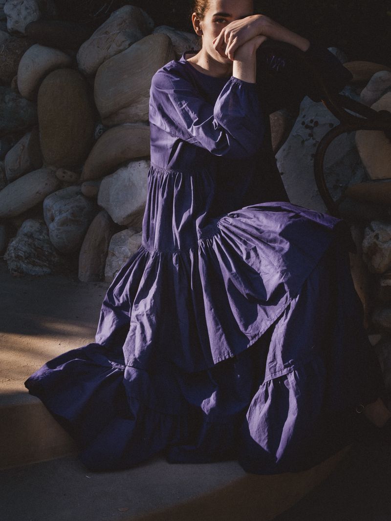 Woman in long purple dress sits with hands clasped near rock wall, creating a contemplative fashion or portrait photograph.