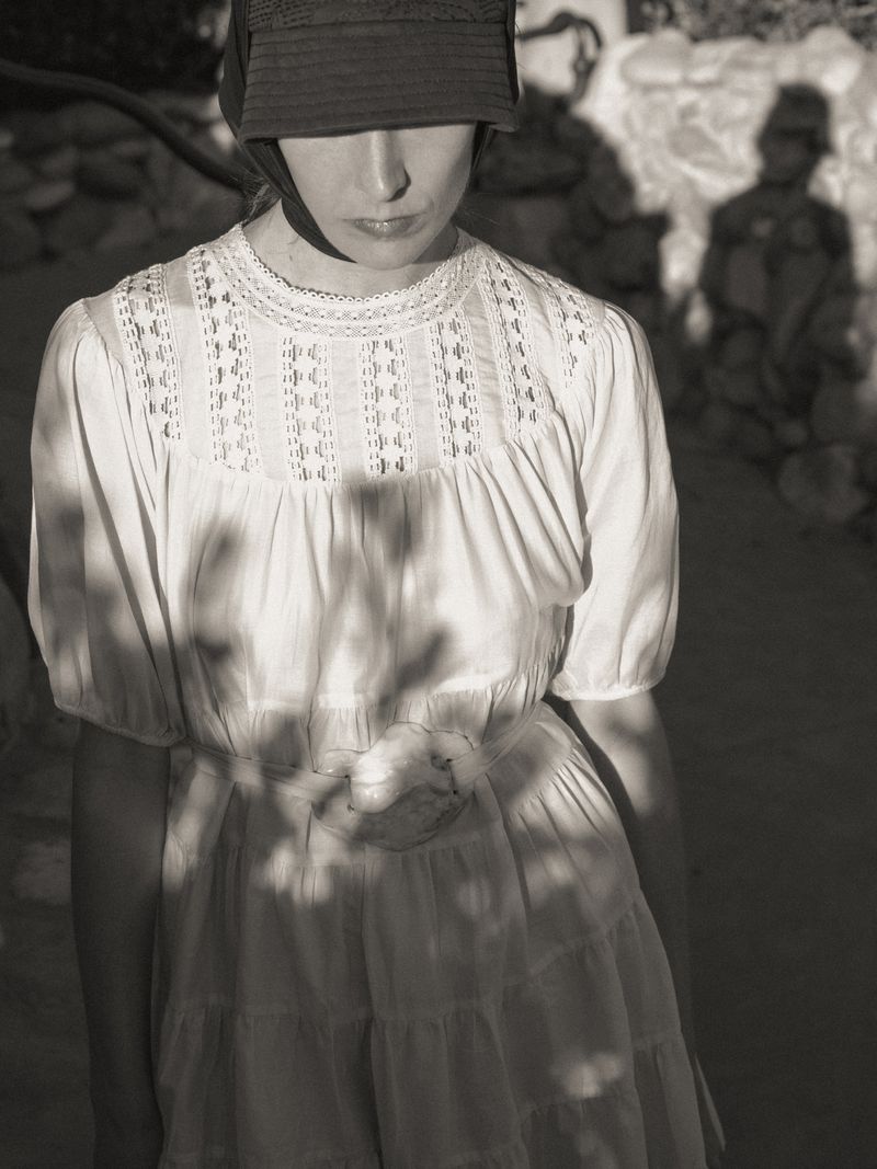 Atherton Grace Johnson in a white lace-trimmed tiered dress and dark brimmed cap under dappled natural sunlight and shadows.