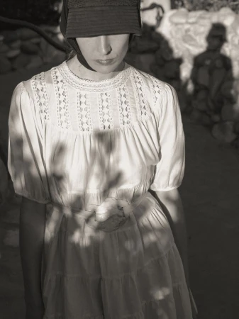 Atherton Grace Johnson in a white lace-trimmed tiered dress and dark brimmed cap under dappled natural sunlight and shadows.