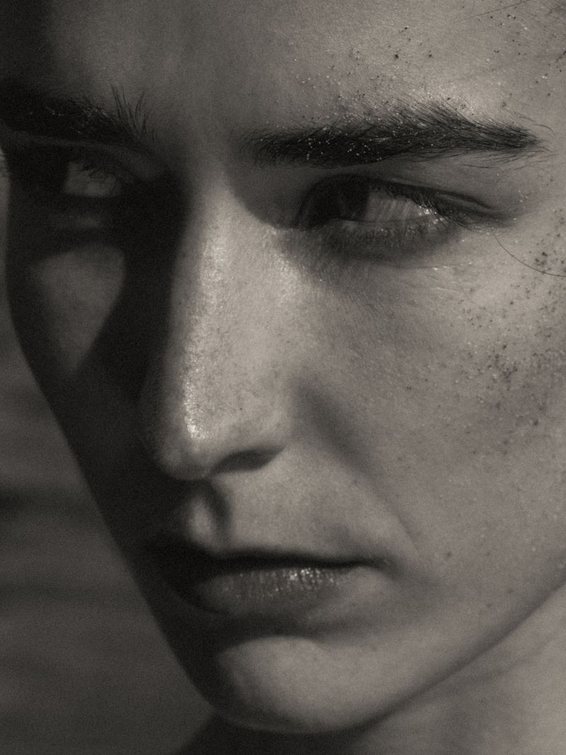 Close-up portrait of a fair-skinned person with subtle makeup, their eyes gazing to the side, set against a neutral gradient background.