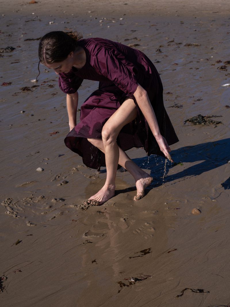 Barefoot woman in a purple dress poses on a sandy beach near the water's edge with overcast light and wet sand.
