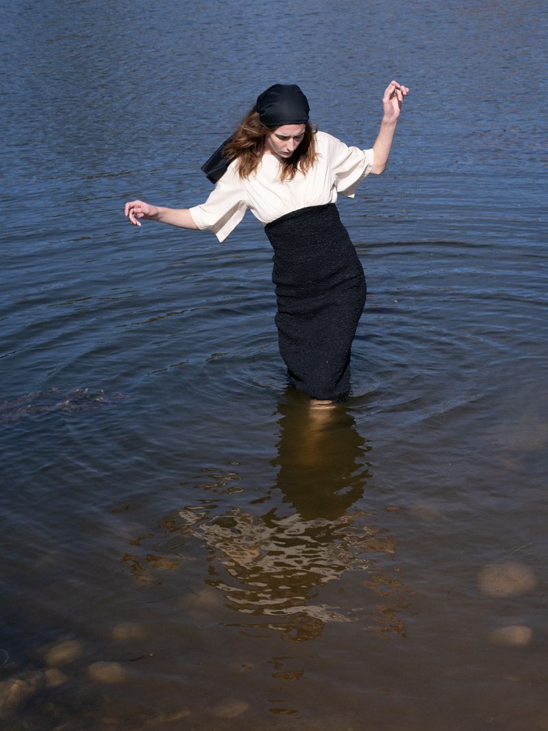 Model Atherton Grace Johnson wades in natural water wearing a cream blouse, black skirt, and a black headscarf.