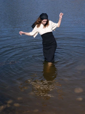 Model Atherton Grace Johnson wades in natural water wearing a cream blouse, black skirt, and a black headscarf.