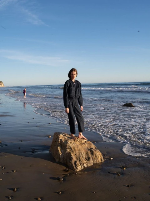 A woman in a navy jumpsuit stands barefoot on a rock at the beach with ocean waves and blue sky in the background in a full shot.