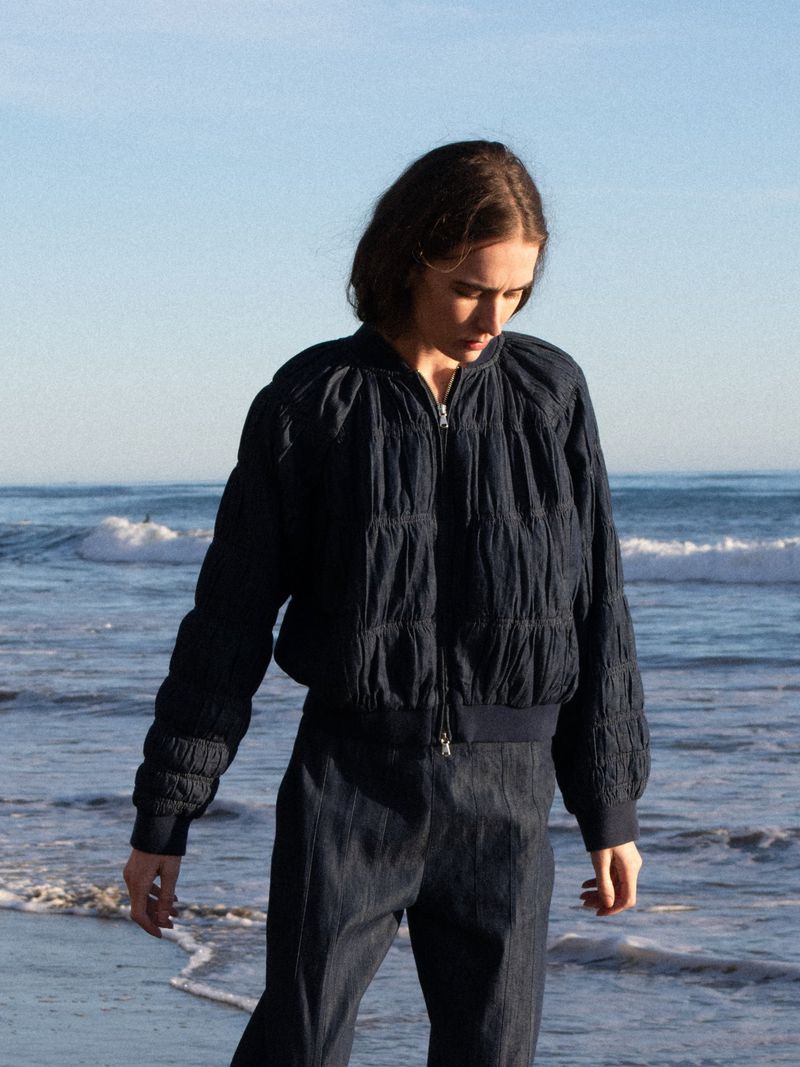 Woman wearing dark blue quilted bomber jacket and trousers on the beach with ocean waves and blue sky in the background.