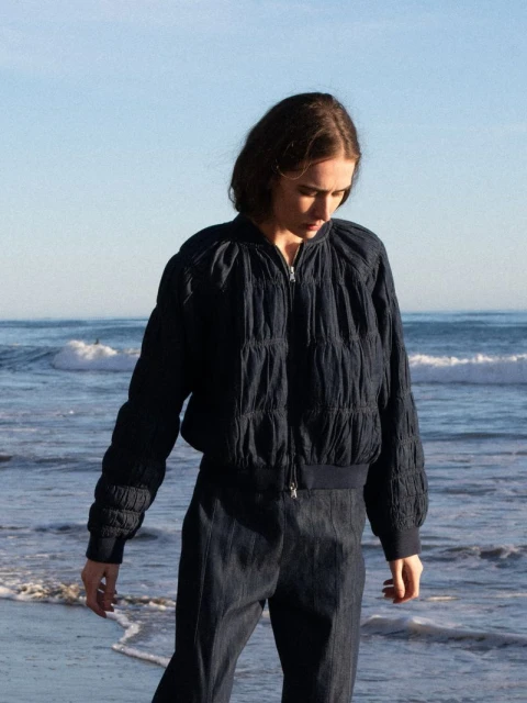 Woman wearing dark blue quilted bomber jacket and trousers on the beach with ocean waves and blue sky in the background.
