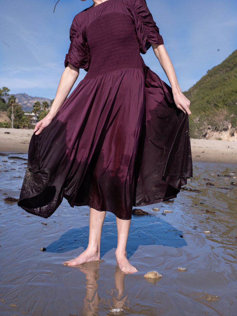 Woman in a burgundy dress stands barefoot on a beach with wet sand, blue sky, and distant hills. Dress appears soaked at the bottom.