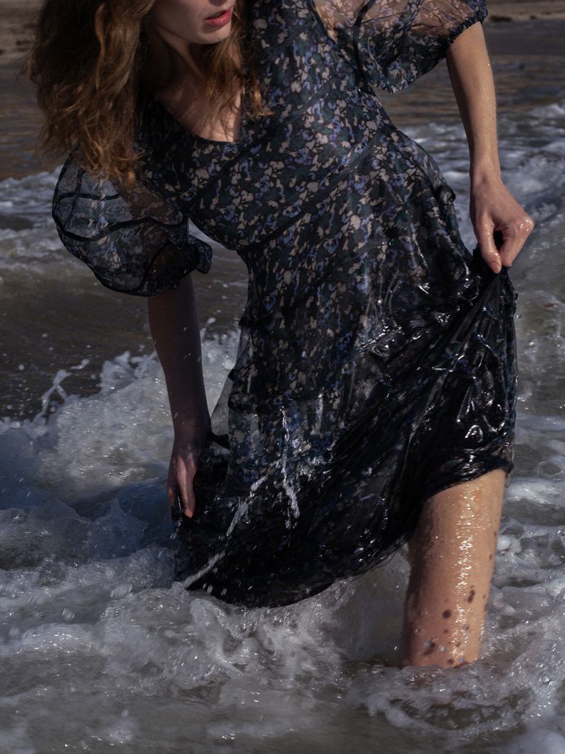 A woman in a floral dress stands in the ocean surf. The water is splashing around her legs, and the dress is partially wet, clinging to her.