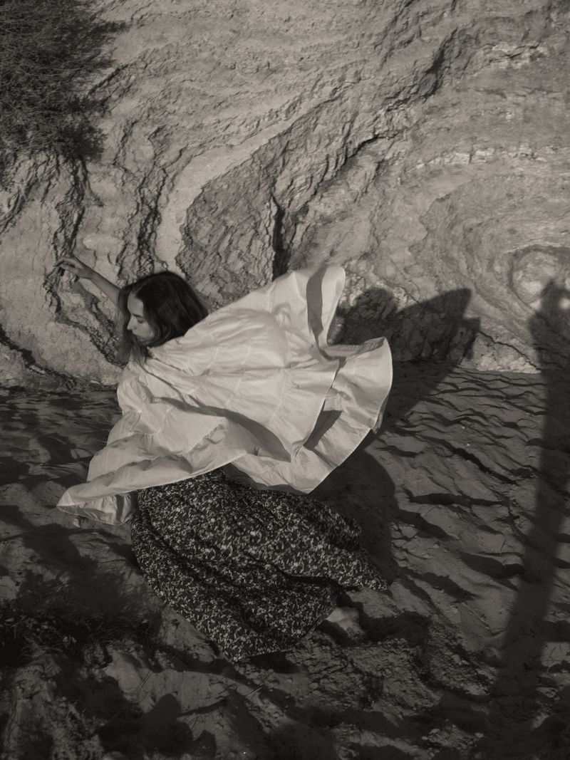 A woman in a light-colored top and patterned skirt poses near a textured rock face in a black and white photograph.