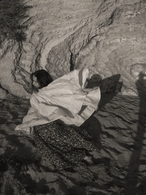 A woman in a light-colored top and patterned skirt poses near a textured rock face in a black and white photograph.