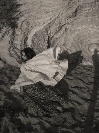 A woman in a light-colored top and patterned skirt poses near a textured rock face in a black and white photograph.