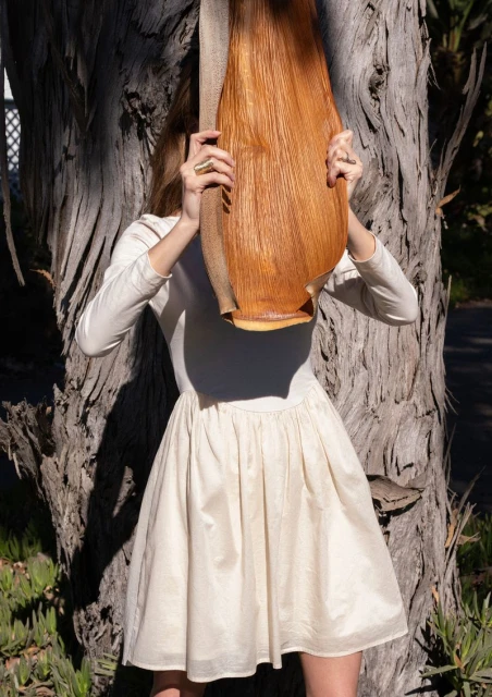 Woman in a cream-colored dress holds a large piece of bark in front of her face outdoors, with a tree trunk behind her.