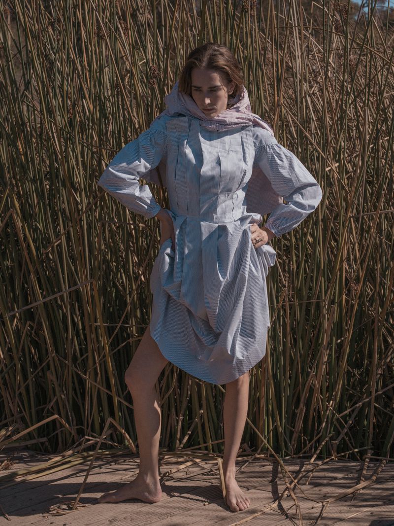 A young woman stands barefoot in a light blue dress and headscarf, posing against tall reeds in a natural outdoor setting on a sunny day.