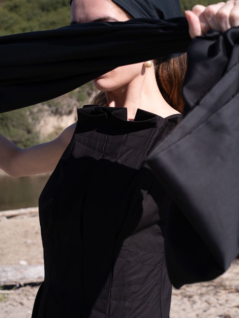 A woman outdoors in a black dress and head covering stands in front of a lake and rocky hills on a sunny day.