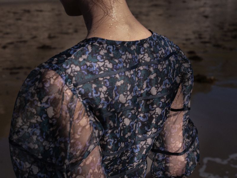 Back view of a woman in a floral dress standing on a beach with dark sand, shallow depth of field, and soft natural lighting.