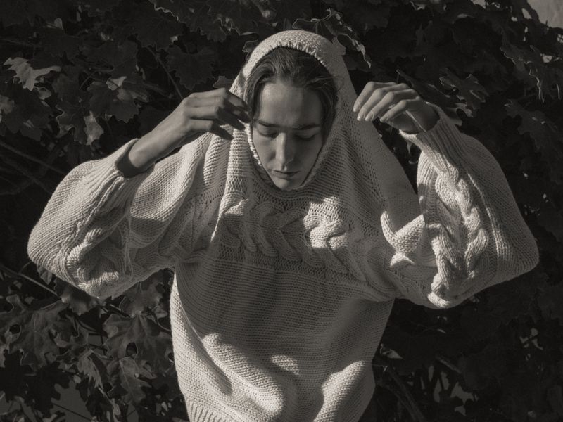 A young adult wearing a cream colored knitted sweater with a hood stands outdoors in front of a leafy foliage background, in a medium shot.