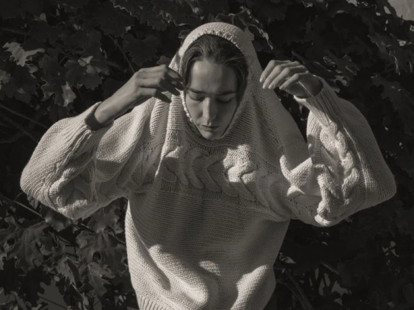A young adult wearing a cream colored knitted sweater with a hood stands outdoors in front of a leafy foliage background, in a medium shot.
