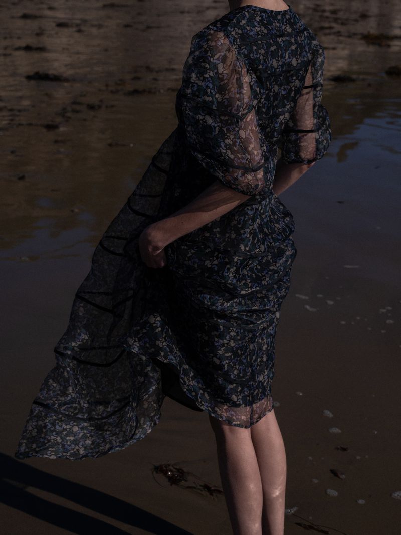 Model wearing a dark sheer patterned puff-sleeve midi dress while standing on wet sand along the shore.