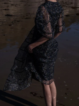 Model wearing a dark sheer patterned puff-sleeve midi dress while standing on wet sand along the shore.