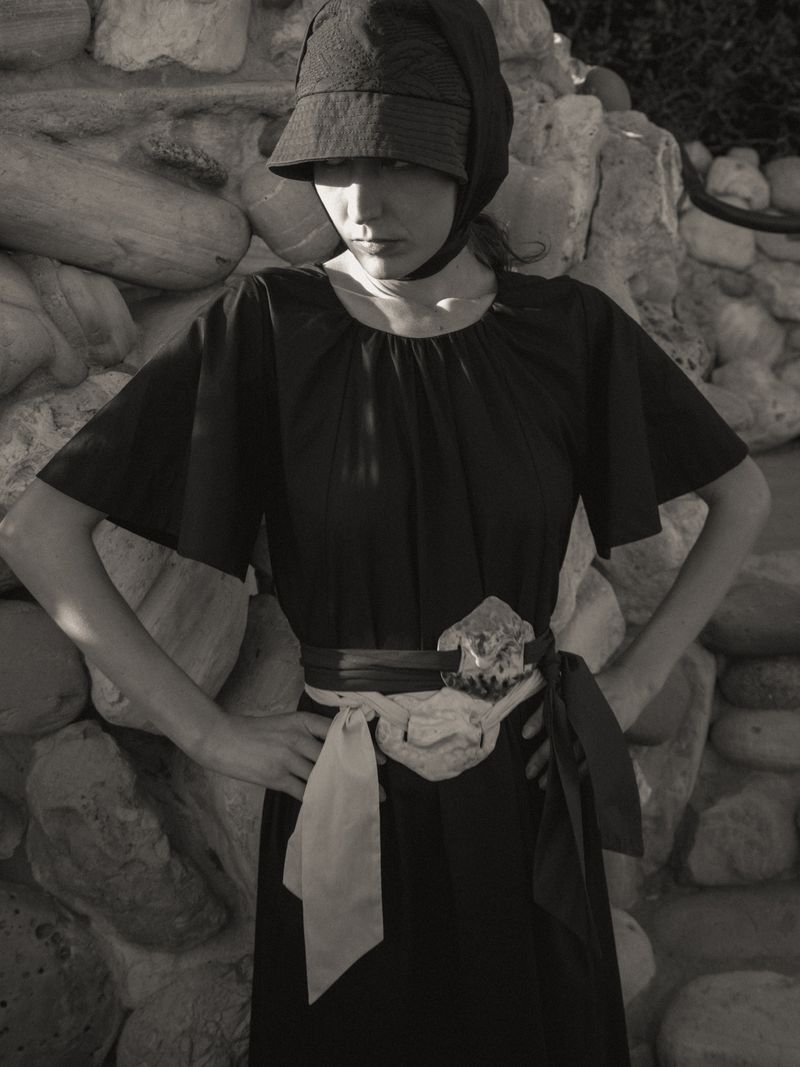 Monochrome portrait of a woman in a vintage-style dress and hat, posing against a stone wall with neutral tones and soft lighting.