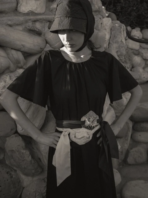 Monochrome portrait of a woman in a vintage-style dress and hat, posing against a stone wall with neutral tones and soft lighting.