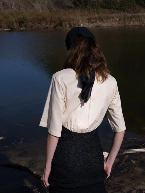 A woman stands near a lake with a cream-colored bolero jacket and a black lace skirt, viewed from the back in a natural setting.