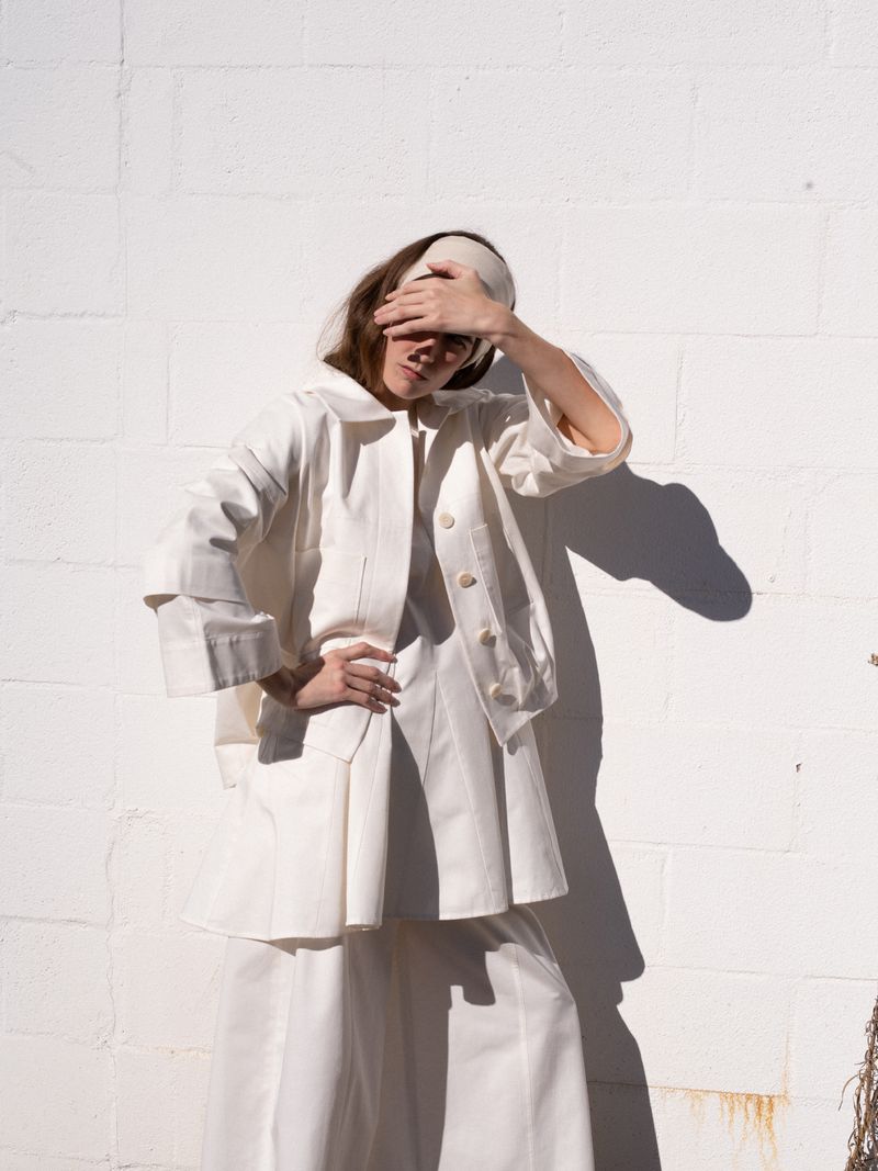 A woman poses in white jacket and wide-leg pants against a textured white wall, her hand partially covering her face in an outdoor setting.