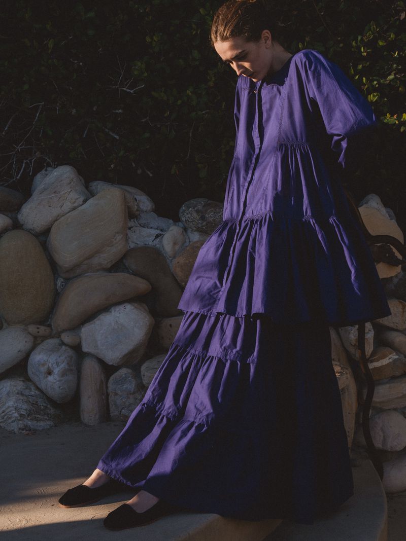 A stylish woman in a tiered purple dress poses near a stone wall in an outdoor fashion shot, showcasing modern design and texture.