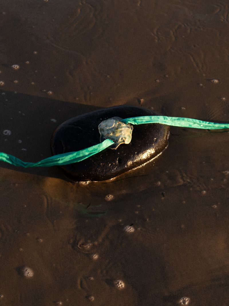 Tethered black stone on wet sand. Turquoise rope is looped through a cloudy clasp. Low-angle view of the beach with no people visible.