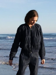Model wearing a dark quilted bomber jacket and matching trousers standing on a sandy beach by the ocean.