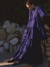 Model wearing a tiered purple dress standing outdoors against a rock wall in natural sunlight for Merlette pre-fall 2025.