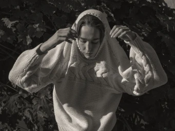 Model Atherton Grace Johnson wearing an oversized cream cable-knit sweater pulled over the head against dark foliage.